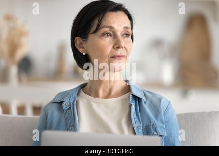 Thoughtful aged woman sitting on sofa looking above notebook screen Stock Photo