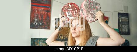 Young caucasian female holding two artistic discs with abstract designs in modern art gallery. Stock Photo