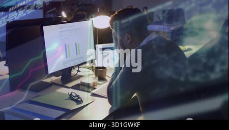 Adult man wearing shirt under sweater viewing charts on screens and tablet in office, with lamp Stock Photo