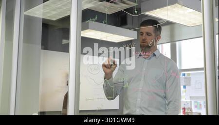 Writing mid adult man in business attire drawing charts on glass partition in office, with marker Stock Photo
