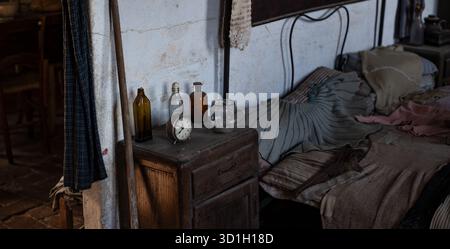 Vintage Bedroom Interior with Antique Alarm Clock and Glass Bottles on Wooden Nightstand Beside Unmade Bed Stock Photo