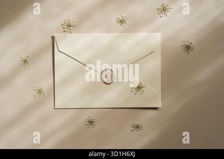 Minimalist flat lay of a white envelope surrounded by tiny dried flowers on a beige background. Elegant stationery mockup with natural light and soft Stock Photo