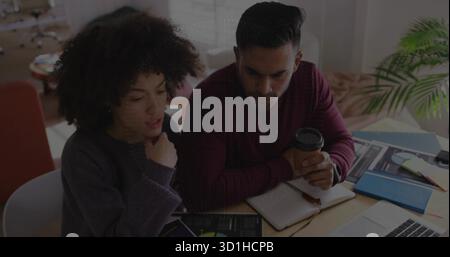 Collaborating coworkers in sweaters reviewing data on tablet and laptop at office, with coffee cup Stock Photo