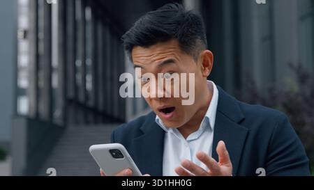 Happy shocked Asian business man looking at phone amazed Korean businessman in city 40s male Chinese Japanese executive feeling surprised wonder Stock Photo