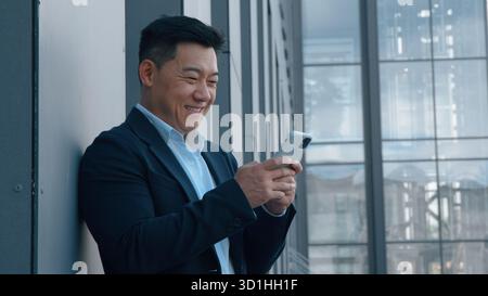 Asian middle-aged business man with mobile phone in city smiling Korean businessman laughing with smartphone reading funny news smile laugh standing Stock Photo