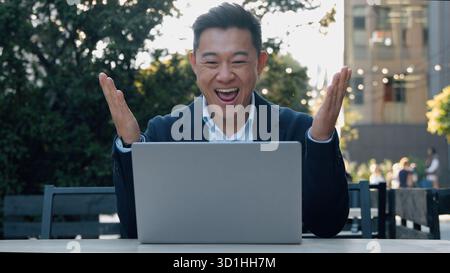 Happy excited Asian business man winning with laptop rejoice looking computer Korean businessman win achieve victory online internet job working in Stock Photo