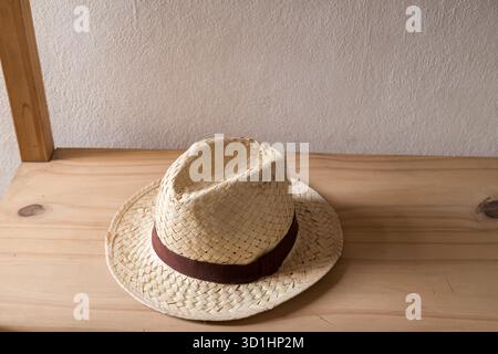 A classic straw fedora hat with a dark brown band rests on a light wooden shelf against a neutral wall. Stock Photo