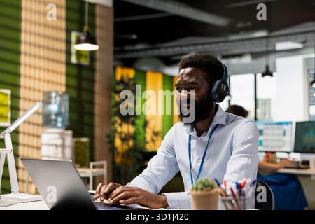 Corporate male employee multitasking with technology, listening to music and reading emails at desk. African american man wearing headphones prepares project presentation on laptop in office. Stock Photo