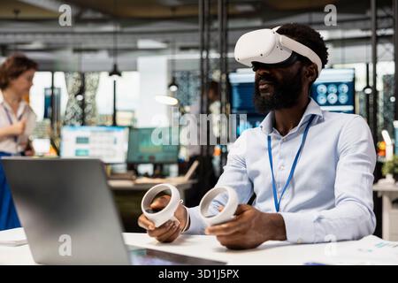 African American executive wears VR headset and holds controllers during interactive meeting on business analytics. Black man financial planning using virtual technology in modern office. Stock Photo