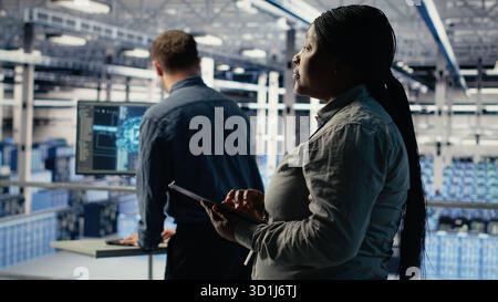 Team of data center technicians checks AI dashboard using laptop during systems review. Teamworking server hub colleagues test artificial intelligence tools on notebook, camera B Stock Photo