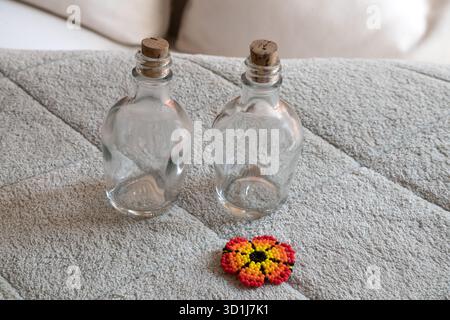 Two empty clear glass bottles with cork stoppers and a vibrant, handmade beaded flower, resting on a textured gray fabric surface. Stock Photo