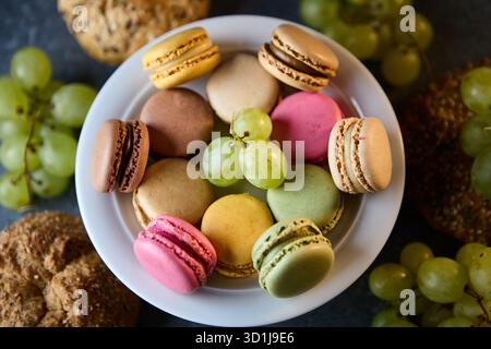 A close up still life showing assorted colorful macarons in a glass bowl surrounded by green and red grapes on a dark stone tabletop. Soft studio ligh Stock Photo