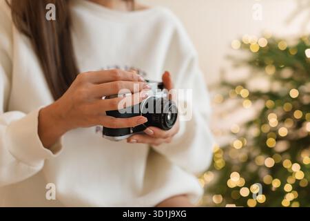 Close-up of a young woman with a camera standing in front of an illuminated Christmas tree in a living room Stock Photo
