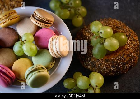 A tight composition featuring pastel macarons alongside a seeded bagel and fresh grapes on a stone surface. Studio lighting highlights texture and col Stock Photo