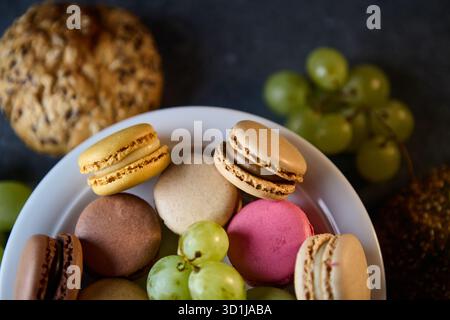 A white bowl filled with assorted macarons rests among bunches of grapes on a dark surface. Shot from above, the scene uses soft light to convey a hom Stock Photo