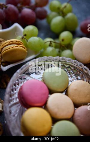 A vertical close up captures colorful macarons in a glass bowl with clusters of grapes behind. The shallow angle and soft light emphasize texture and Stock Photo