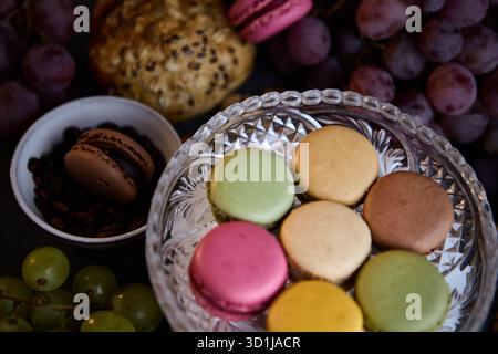 A cozy tabletop scene features a crystal bowl of macarons surrounded by grapes, a cookie and a small dish. The soft light and warm colors create an in Stock Photo