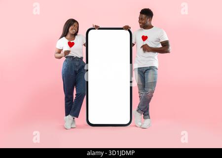 Couple Holds Paper Hearts While Standing Next to a Big Smartphone With an Empty Screen Stock Photo