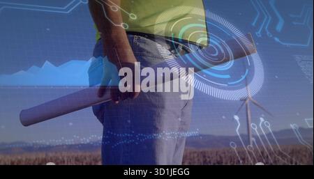 Standing engineer holding blueprint in farm field at sunset, with HUD overlays and wind turbine Stock Photo