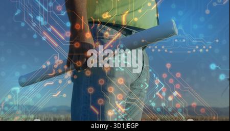 Standing man wearing yellow shirt holding blueprint at hip on rooftop, with digital circuit overlay Stock Photo