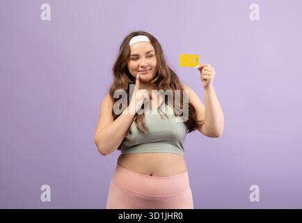 A Pensive Plus Size European Millennial Female in Sporty Attire Smiles While Holding a Credit Card Stock Photo
