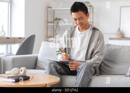 Young Asian man writing something in notebook on sofa at home Stock Photo