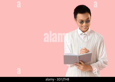 Young Asian man writing something in notebook on pink background Stock Photo