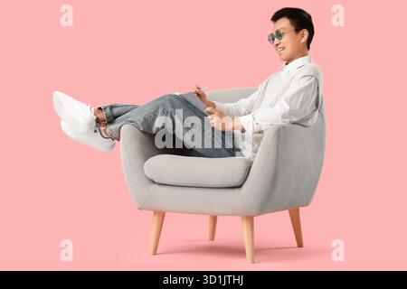 Young Asian man writing something in notebook on armchair against pink background Stock Photo