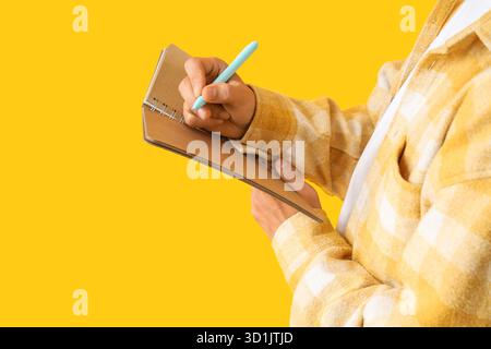 Young man writing something in notebook on yellow background, closeup Stock Photo