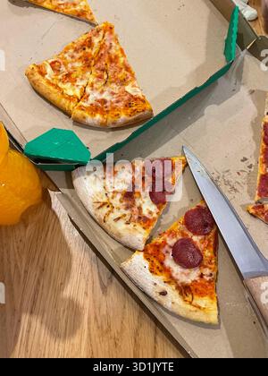 Close-up of leftover pizza slices in a cardboard delivery box on a wooden table. Slices topped with pepperoni sausage and cheese. A knife lying nearby Stock Photo