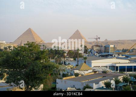 View of the Giza Pyramids rising behind a modern urban area with palm trees, buildings, and construction cranes in the foreground Stock Photo