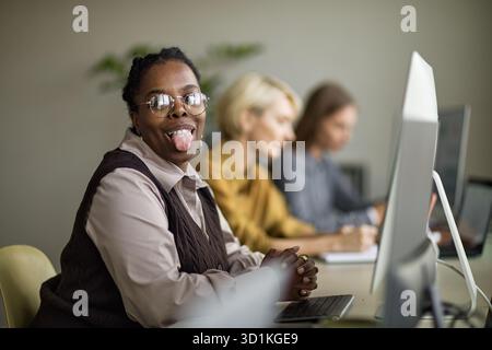 Portrait of Positive Young Adult Black Woman Sticking out Tongue at Office Desk Stock Photo