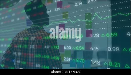 Studying analyst in plaid shirt reviewing stock charts on glass wall in office, with desk behind Stock Photo