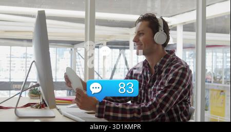 Smiling man in plaid shirt, headphones holding coffee mug, viewing monitor in office with succulent Stock Photo