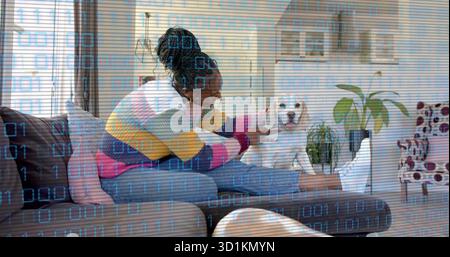 Leaning woman in multicolored sweater and sneakers petting white puppy on gray sofa, with plant Stock Photo