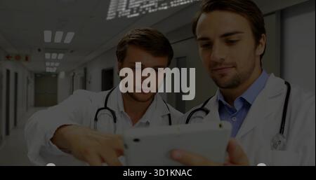 Reviewing doctors wearing lab coats and stethoscopes consulting on tablet in hospital corridor Stock Photo