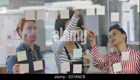Three professionals collaborating behind office glass board, with colorful sticky notes and markers Stock Photo