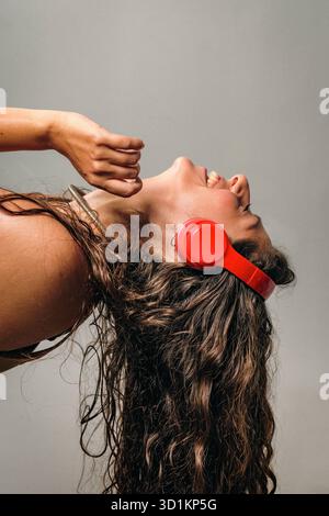 A young woman laughs joyfully while wearing bright red headphones. She tilts her head back, letting her long, curly hair cascade as she loses herself Stock Photo
