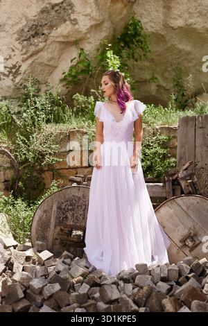 young woman in a long white dress poses elegantly among rocks and greenery, showcasing both her style and the beauty of nature in the background under Stock Photo
