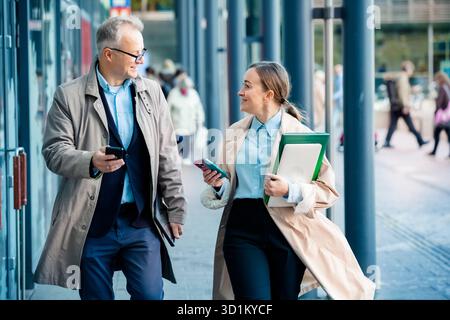 Successful business man and woman walking in city street, smiling and talking with laptop and coffee. Modern urban teamwork and success concept. Stock Photo