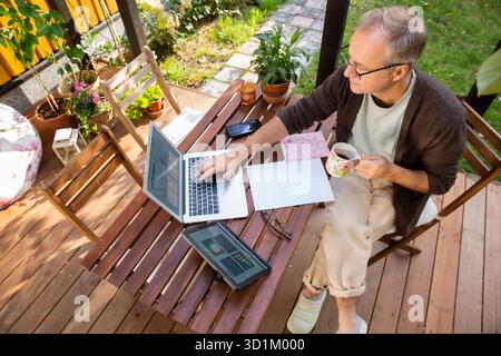 Middle aged man working remotely on laptop at cozy sunny patio, digital nomad lifestyle, outdoor home office, freelance and productivity concept Stock Photo