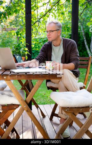 Middle aged man working remotely on laptop at cozy sunny patio, digital nomad lifestyle, outdoor home office, freelance and productivity concept Stock Photo