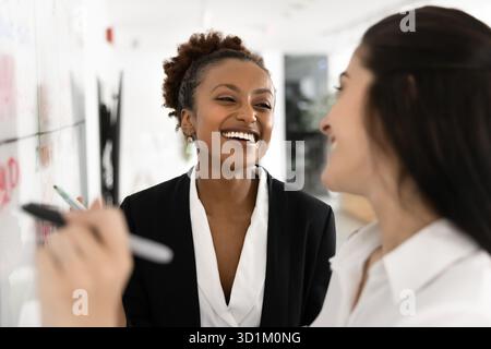 Two laughing young diverse women writing on glass schedule board Stock Photo