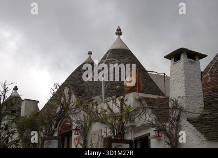 A close view of traditional trulli houses with conical roofs made of layered gray stone. One roof bears a white painted symbol, while whitewashed wall Stock Photo