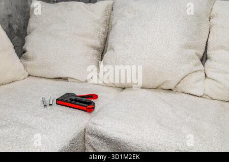 Close up of a black upholstery stapler and metal staples placed on a wooden frame covered with soft white fabric. Concept of furniture renovation, DIY Stock Photo