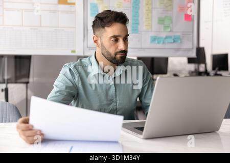 Young male entrepreneur working at desk on notebook holding papers Stock Photo
