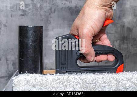 Close up of a black upholstery stapler and metal staples placed on a wooden frame covered with soft white fabric. Concept of furniture renovation, DIY Stock Photo