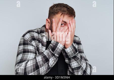 Man in plaid shirt covering face with hands, peeking through fingers. Stock Photo