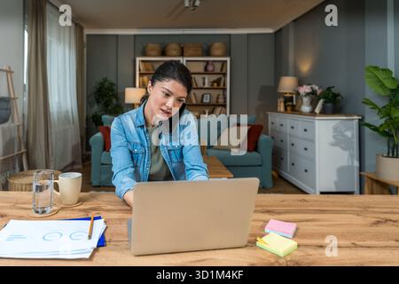 Freelance businesswoman in home office reviewing charts and dictating a voice note while analyzing financial reports on a laptop — remote work, strate Stock Photo