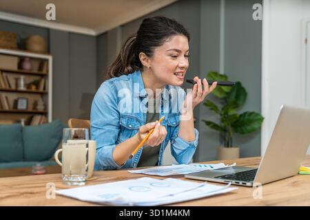 Freelance businesswoman in home office reviewing charts and dictating a voice note while analyzing financial reports on a laptop — remote work, strate Stock Photo